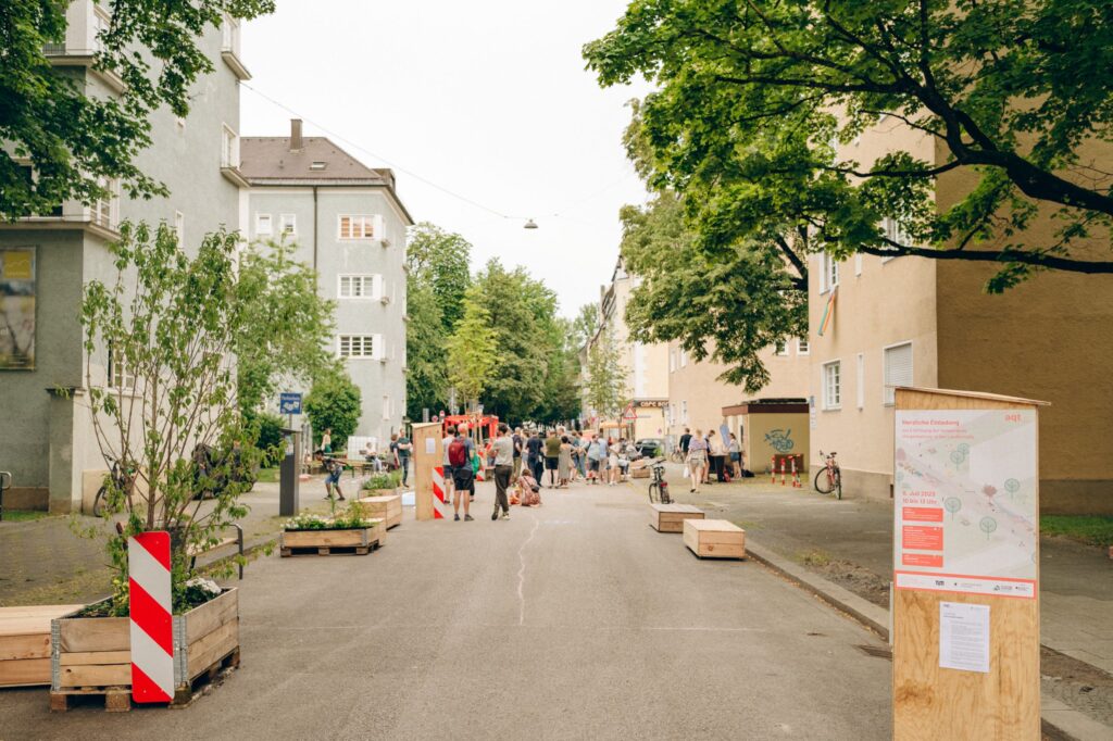 Das Bild zeigt eine belebte Straße, die offensichtlich für den Verkehr gesperrt und in einen öffentlichen Begegnungsraum verwandelt wurde. Im Vordergrund sind Holzkisten mit Bepflanzung und Sitzbänke auf der Straße platziert. Eine Gruppe von Menschen versammelt sich weiter hinten, wahrscheinlich bei einer Veranstaltung oder einem Straßenfest. Die Umgebung wirkt freundlich und einladend, mit viel Grün und Wohngebäuden auf beiden Seiten.